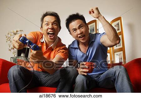 Stock Photograph - Two men sitting side by side, one man holding game controller, the other raising hand in a fist. Fotosearch