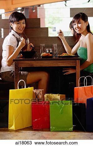 Stock Image - Two young women sitting at table in restaurant, surrounded by shopping bags, smiling at camera. Fotosearch