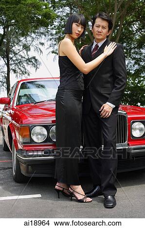 Well dressed couple standing in front of red car, woman with hands on man's shoulders View Large Photo Image Picture - Well dressed couple standing in front of red car, woman with hands on man's shoulders. Fotosearch