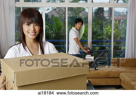 Woman carrying a box, smiling at camera, man in the background View Large Photo Image Picture - Woman carrying a box, smiling at camera, man in the background. Fotosearch