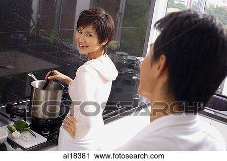 Stock Image - Woman cooking on stove, looking over shoulder at man standing behind her. Fotosearch