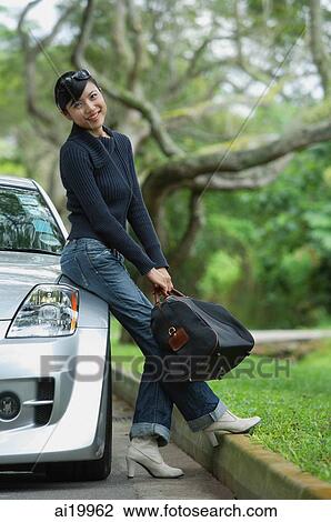 Stock Image - Woman holding bag, leaning on car, smiling at camera. Fotosearch