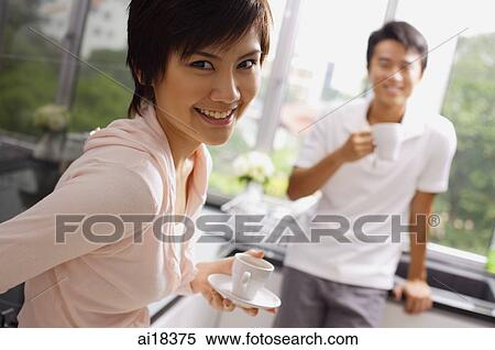 Stock Photography - Woman holding cup and saucer, smiling at camera, man in the background. Fotosearch