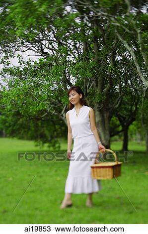 Stock Image - Woman holding picnic basket, walking in park. Fotosearch