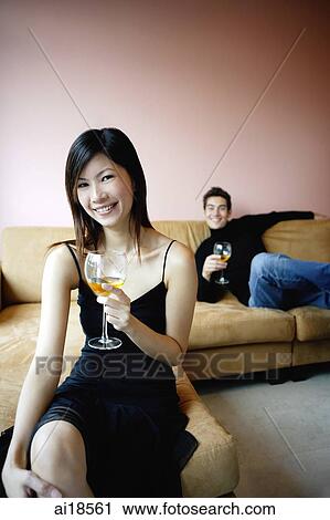 Stock Image - Woman in black dress holding wine glass, smiling at camera, man in the background. Fotosearch