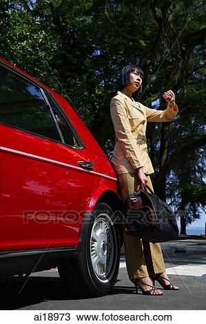 Stock Image - Woman leaning on red car, carrying bag, looking at watch. Fotosearch
