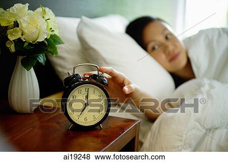 Woman lying in bed, reaching for alarm clock on bedside table View Large Photo Image Stock Photo - Woman lying in bed, reaching for alarm clock on bedside table. Fotosearch