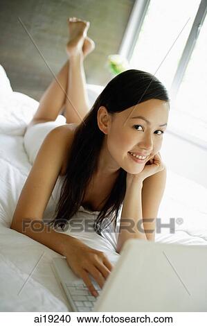 Stock Image - Woman lying on bed, using laptop, smiling at camera. Fotosearch