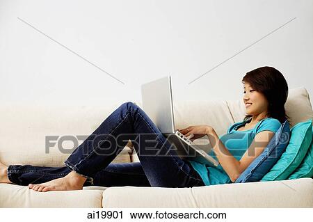 Stock Image - Woman lying on sofa, using laptop. Fotosearch