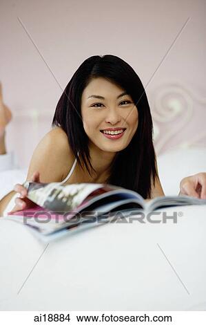 Picture - Woman on bed, flipping through magazine, smiling at camera. Fotosearch