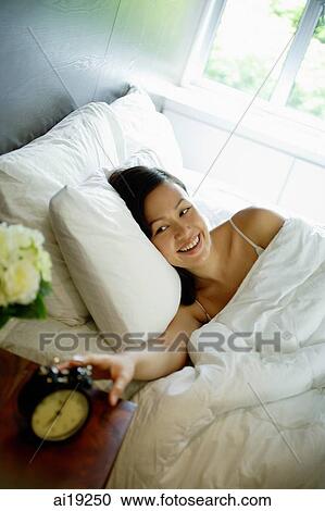 Stock Image - Woman reaching for alarm clock on bedside table, smiling. Fotosearch