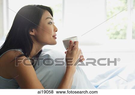 Woman sitting on bed, holding cup, looking away View Large Photo Image Stock Photo - Woman sitting on bed, holding cup, looking away. Fotosearch