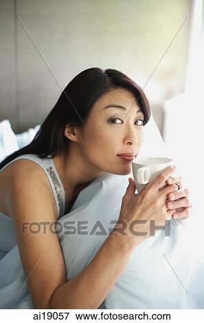 Woman sitting on bed, holding cup to lips, looking at camera View Large Photo Image Stock Photo - Woman sitting on bed, holding cup to lips, looking at camera. Fotosearch