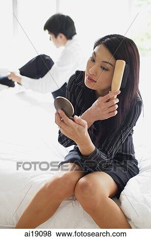 Woman sitting on bed, looking at compact, brushing hair, man in the background View Large Photo Image Stock Photo - Woman sitting on bed, looking at compact, brushing hair, man in the background. Fotosearch
