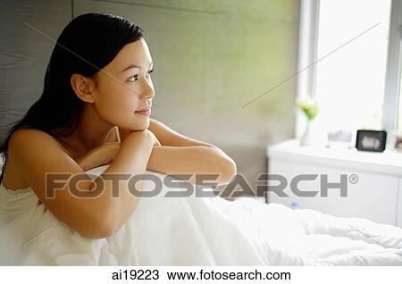 Stock Image - Woman sitting on bed, resting head on knees, looking away. Fotosearch