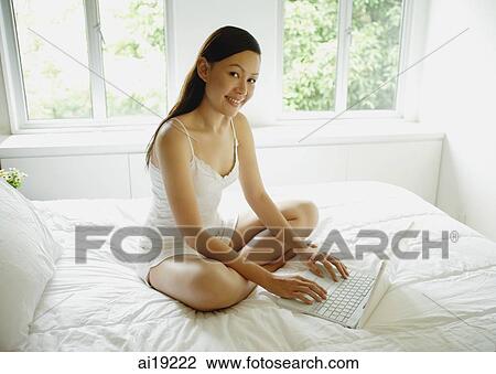 Woman sitting on bed, using laptop, smiling at camera View Large Photo Image Stock Image - Woman sitting on bed, using laptop, smiling at camera. Fotosearch