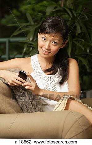 Woman sitting on sofa, holding mobile phone, looking at camera View Large Photo Image Stock Image - Woman sitting on sofa, holding mobile phone, looking at camera. Fotosearch