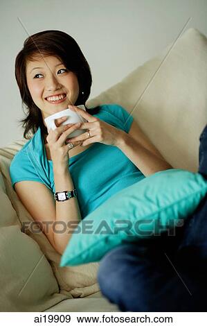 Stock Photo - Woman sitting on sofa, holding mug, looking away. Fotosearch
