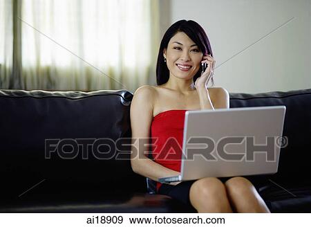 Stock Photo - Woman sitting on sofa, using laptop and mobile phone, smiling at camera. Fotosearch