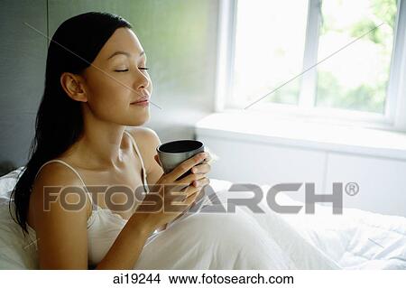 Woman sitting up in bed, holding cup, eyes closed View Large Photo Image Picture - Woman sitting up in bed, holding cup, eyes closed. Fotosearch