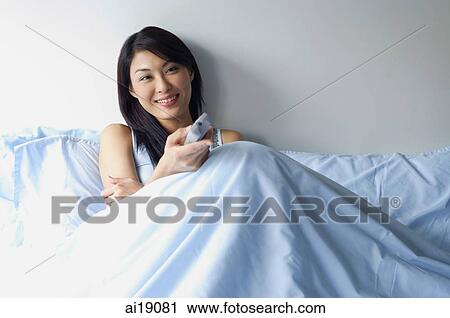 Woman sitting up in bed, holding TV remote control, watching TV View Large Photo Image Stock Image - Woman sitting up in bed, holding TV remote control, watching TV. Fotosearch