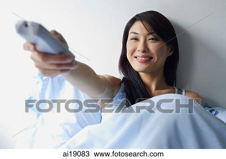 Stock Image - Woman sitting up in bed, holding TV remote control, arm outstretched. Fotosearch