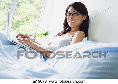 Woman sitting up on bed using laptop, looking at camera View Large Photo Image Stock Photo - Woman sitting up on bed using laptop, looking at camera. Fotosearch