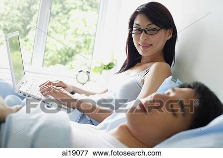 Woman sitting up on bed with laptop, looking at man sleeping next to her View Large Photo Image Stock Photo - Woman sitting up on bed with laptop, looking at man sleeping next to her. Fotosearch