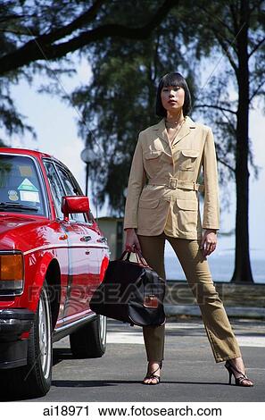 Stock Image - Woman standing by car, carrying bag. Fotosearch