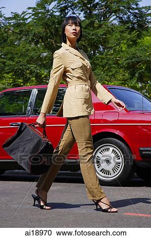 Stock Image - Woman walking by red car, carrying bag. Fotosearch
