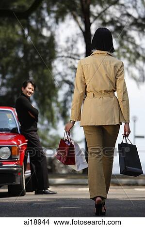 Woman with shopping bags, walking towards man standing by car, rear view View Large Photo Image Picture - Woman with shopping bags, walking towards man standing by car, rear view. Fotosearch