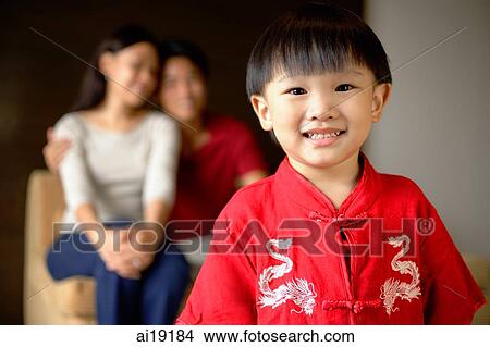 Picture - Young boy in red shirt smiling at camera, parents in background. Fotosearch