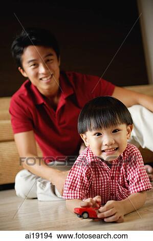 Young boy lying on floor, holding toy car, father sitting behind him View Large Photo Image Picture - Young boy lying on floor, holding toy car, father sitting behind him. Fotosearch
