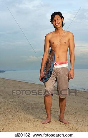 Stock Image - Young man holding skimboard, standing on beach. Fotosearch