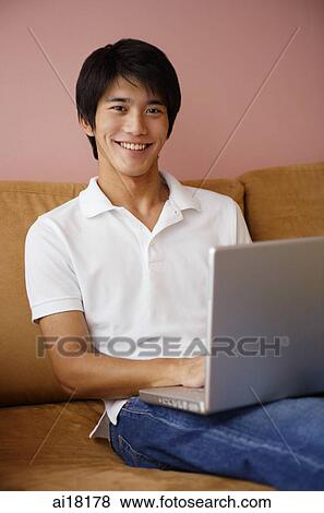 Young man on sofa, using laptop, smiling at camera View Large Photo Image Stock Photo - Young man on sofa, using laptop, smiling at camera. Fotosearch