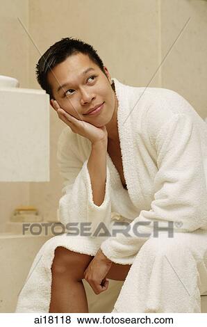 Stock Photo - Young man sitting in bathroom, hand on chin, looking away. Fotosearch