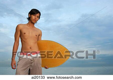 Stock Image - Young man with skimboard, looking at camera. Fotosearch