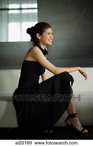 Stock Photo - Young woman in black dress, crouching, looking away. Fotosearch