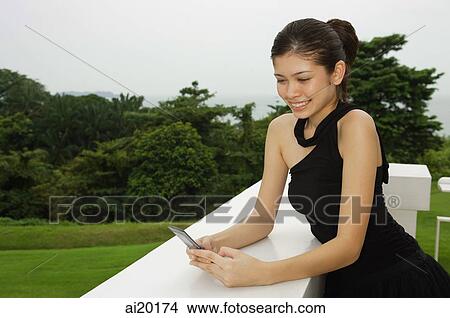 Young woman in black dress, leaning on ledge, looking at mobile phone View Large Photo Image Picture - Young woman in black dress, leaning on ledge, looking at mobile phone. Fotosearch