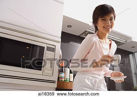 Young woman in kitchen, holding cup and saucer, smiling View Large Photo Image Stock Photo - Young woman in kitchen, holding cup and saucer, smiling. Fotosearch