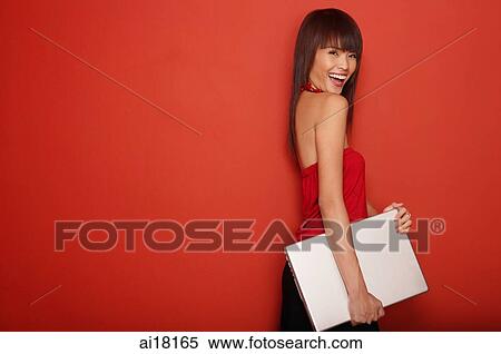Stock Photography - Young woman in red tube top, standing against red wall, carrying laptop. Fotosearch
