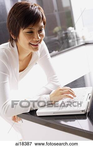 Stock Photo - Young woman leaning on kitchen counter, using laptop. Fotosearch