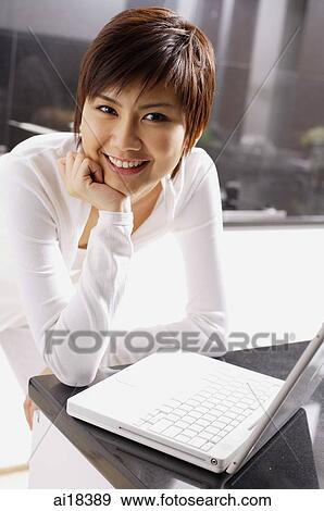 Stock Photo - Young woman leaning on kitchen counter, hand on chin, laptop open in front of her. Fotosearch