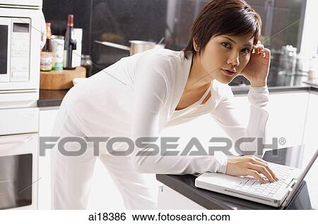 Young woman leaning on kitchen counter, using laptop, looking at camera View Large Photo Image Stock Photograph - Young woman leaning on kitchen counter, using laptop, looking at camera. Fotosearch
