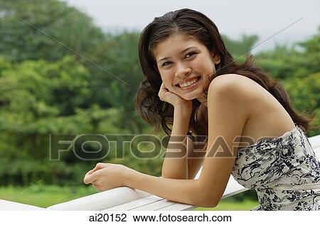 Young woman leaning on railing, hand on chin, smiling at camera View Large Photo Image Stock Image - Young woman leaning on railing, hand on chin, smiling at camera. Fotosearch