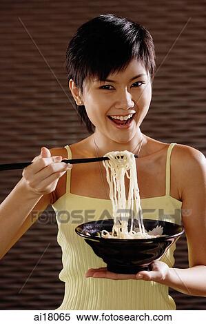 Stock Photography - Young woman lifting noodles from bowl with chopsticks, smiling at camera. Fotosearch