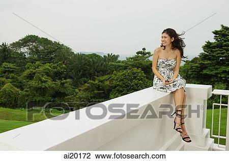 Stock Image - Young woman sitting on balcony ledge, looking away. Fotosearch