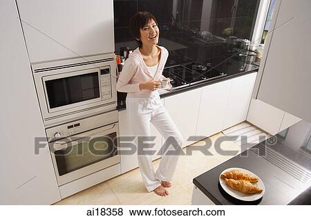 Stock Photo - Young woman standing in kitchen, holding cup and saucer, smiling at camera. Fotosearch