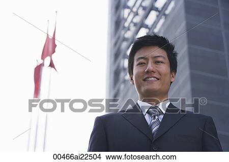 Picture - A businessman looks at the camera as he stands in front of a building with flags. Fotosearch
