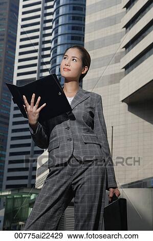 Stock Photo - A businesswoman holds open a folder as she stands in front of skyscrapers. Fotosearch